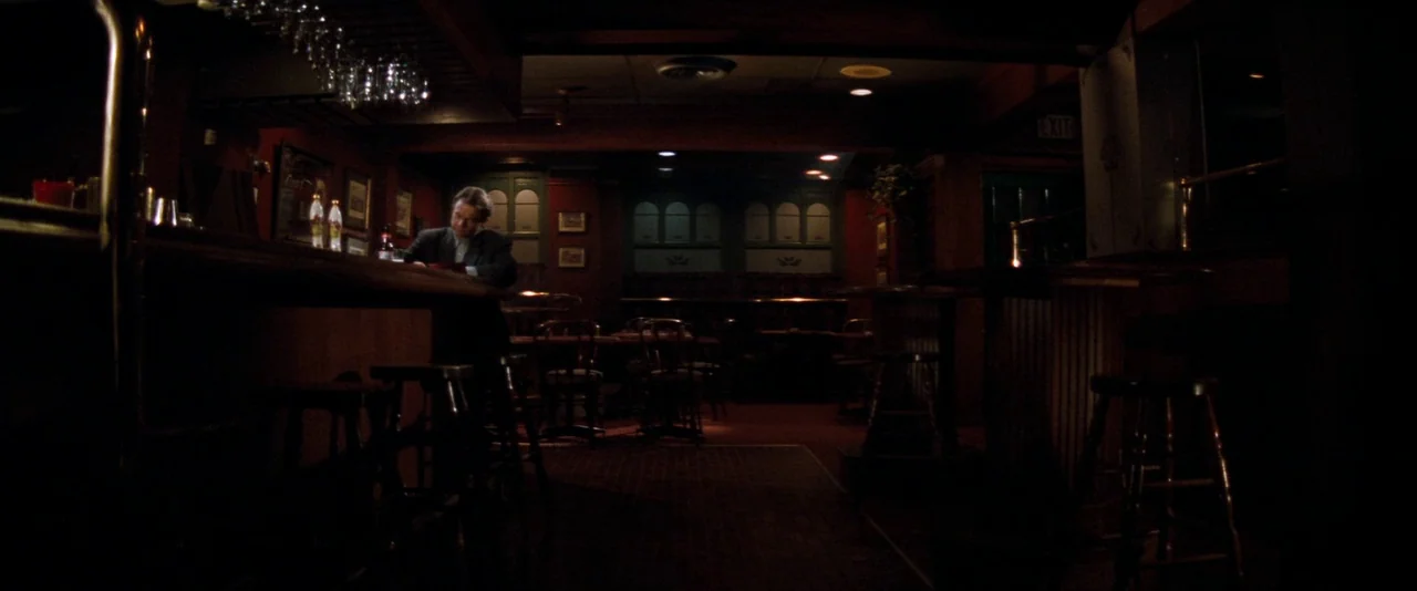 In The Mouth of Madness (1994) movie still: wide — A dark empty bar with wood paneling and numerous wooden stools arranged around tables. A lone man…