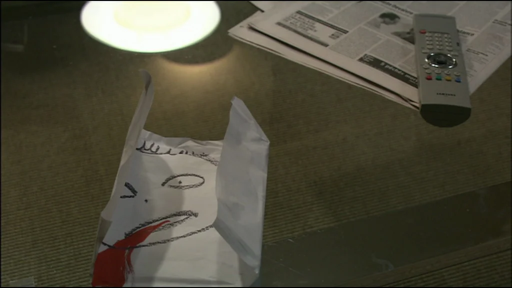 Caché (Hidden) (2005) movie still: closeup — A glass coffee table holds a remote, a newspaper, and a folded paper drawing. The drawing features a…