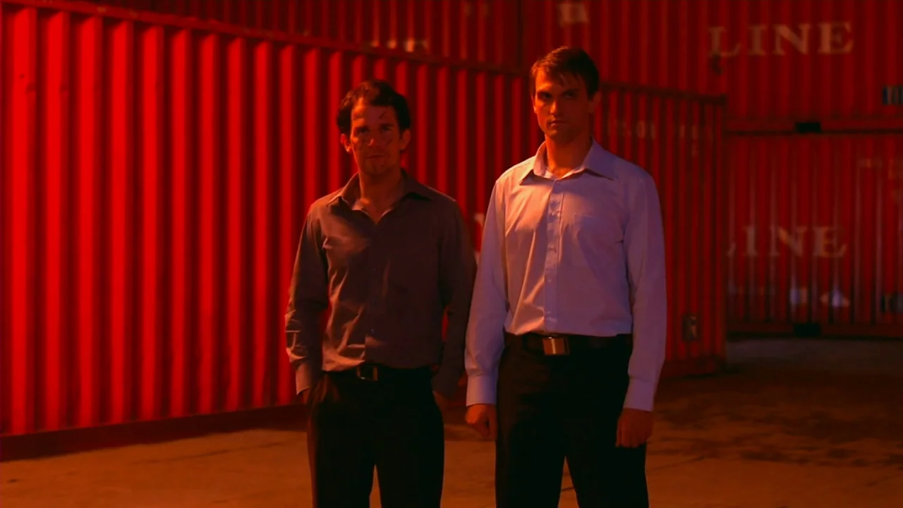 Merantau (2009) movie still: fullbody — A dark outdoor shipping area surrounded by tall red corrugated metal shipping containers at night. Two men…