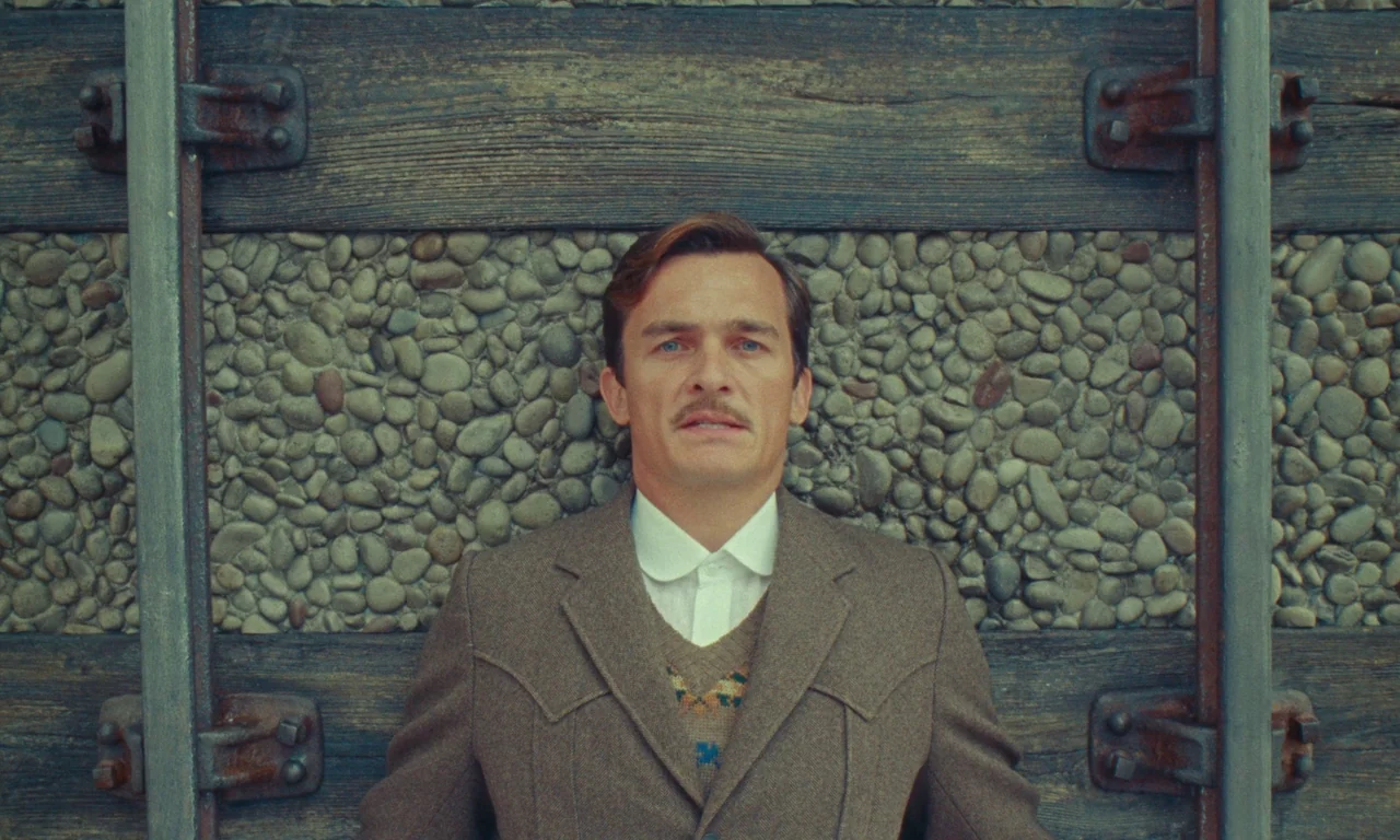 The Swan (2023) movie still: medium — A man stands centered before a rustic retaining wall built of timber and pebbles. The textured stone and wood…