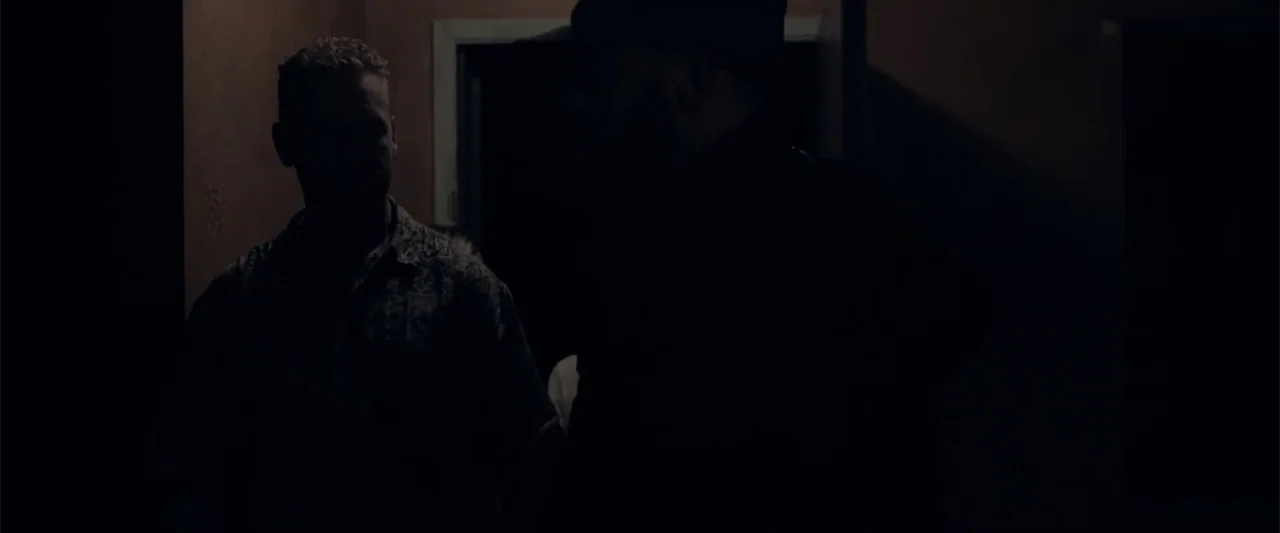 Anomaly (2006) movie still: medium — A dimly lit interior space with dark walls and a doorway in the background. Two men stand in the shadows facing…