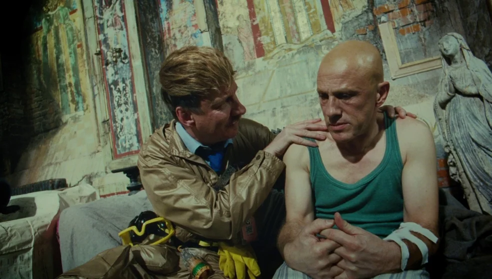 The Zero Theorem (2013) movie still: medium — A dilapidated interior space features faded frescoes on crumbling walls and a stone statue. A man in a…