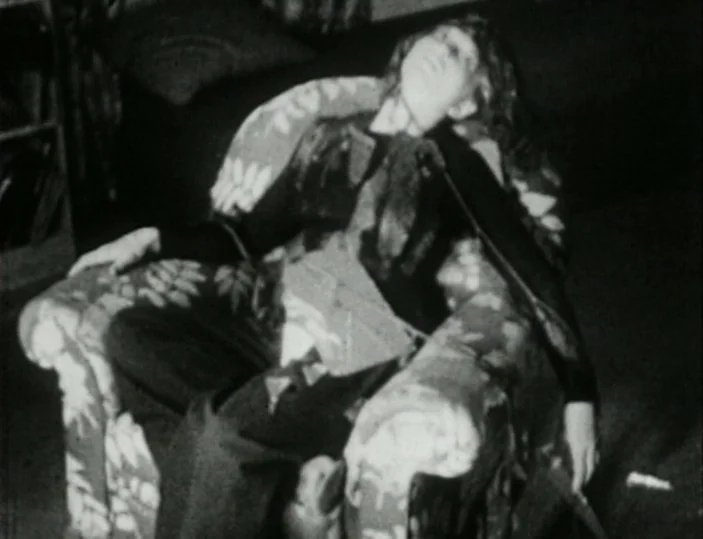 Meshes of the Afternoon (1943) movie still: medium — A person sits in a patterned fabric armchair in a dimly lit room. The individual rests their…