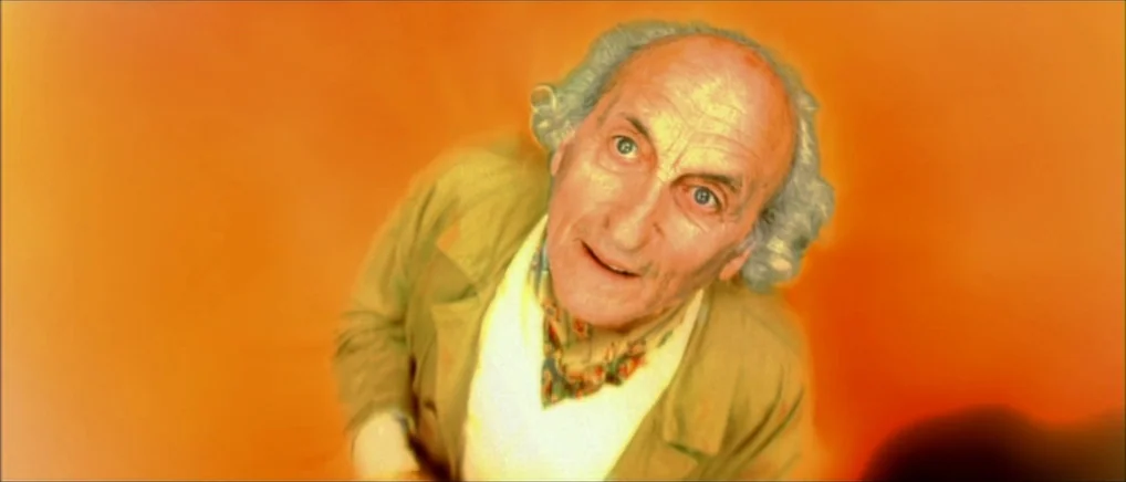 Amelie (2001) movie still: closeup — A man against a solid orange background looks upward with a direct gaze. His head and torso are positioned…