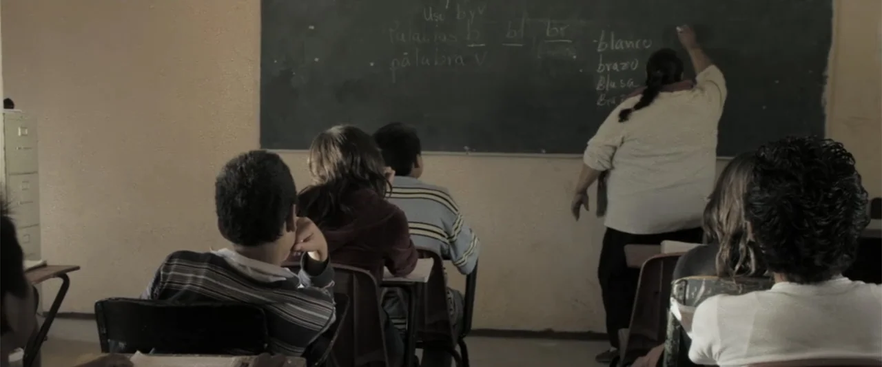 At Dawn (2013) movie still: medium — A classroom with desks arranged in rows facing a dark slate blackboard wall. An adult female teacher stands at…