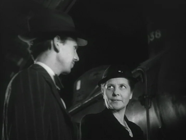 Crisis (1946) movie still: medium — A dark train station platform at night with a large locomotive partially visible. Two people stand close…