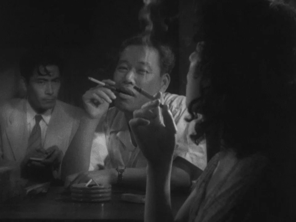 Stray Dog (1949) movie still: medium — A dark, dimly lit environment features a wooden table with an ashtray and cigarettes. A man holds a cigarette…
