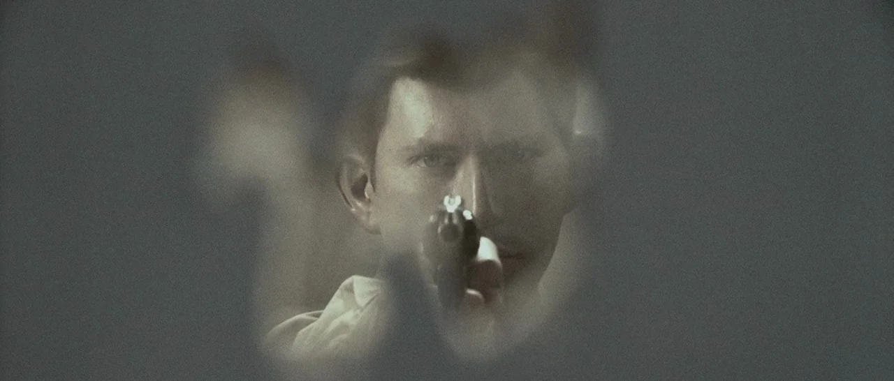 Slice (2018) movie still: closeup — A man holds a handgun directly toward the viewer from a dark, indistinct location. His face is framed by a soft,…