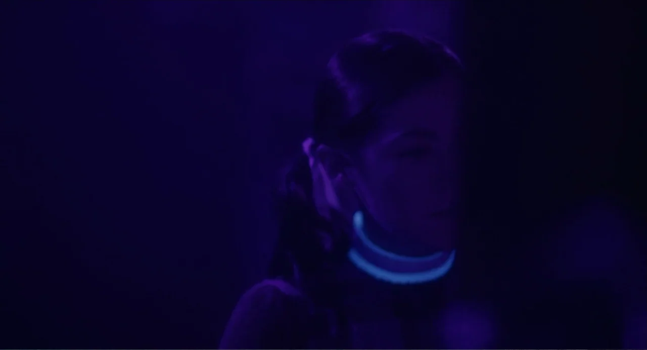 Orphan: First Kill (2022) movie still: closeup — A dark space lit by pervasive ultraviolet purple light. A young woman stands with a glowing cyan…