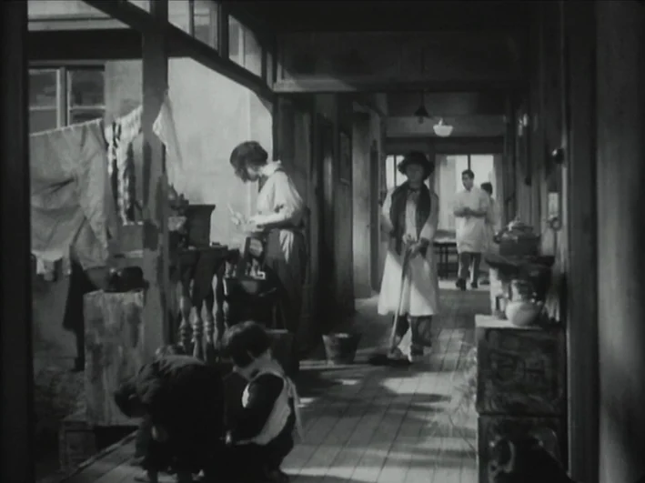 The Quiet Duel (1949) movie still: wide — A narrow residential corridor contains wooden floorboards and laundry hanging on a rack. A woman stands in…