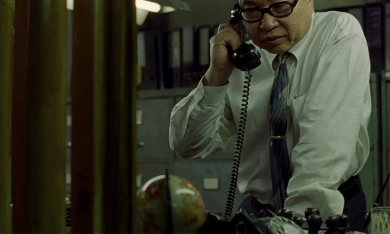 In the Mood for Love (2000) movie still: medium — A cluttered office filled with metal filing cabinets under low, dim fluorescent light. A man in a…