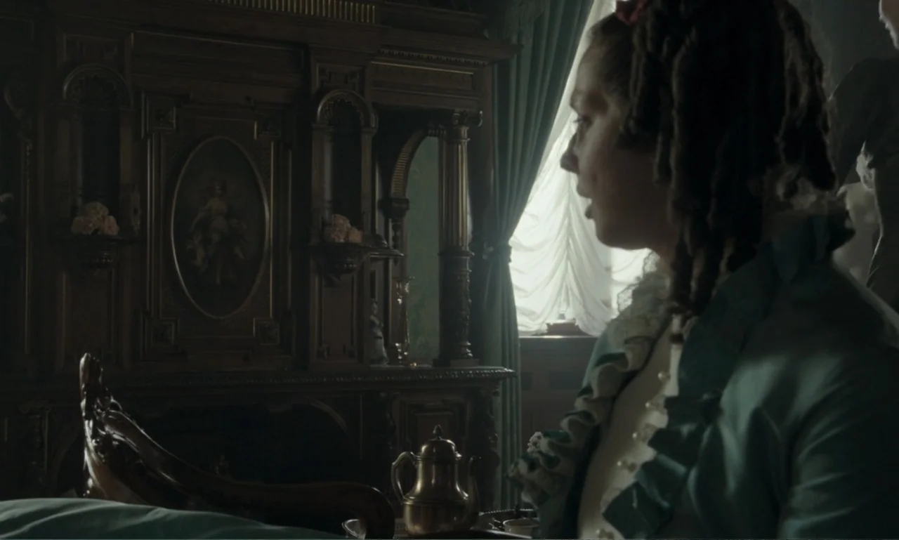 The Ugly Stepsister (2025) movie still: medium — A historic interior features an ornate dark wood wardrobe and a sunlit window. A woman in a teal…