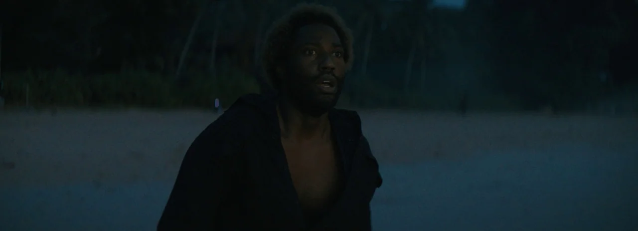 The Creator (2023) movie still: medium — A dark, quiet beach at night features tall silhouettes of palm trees nearby. A man with an afro and…