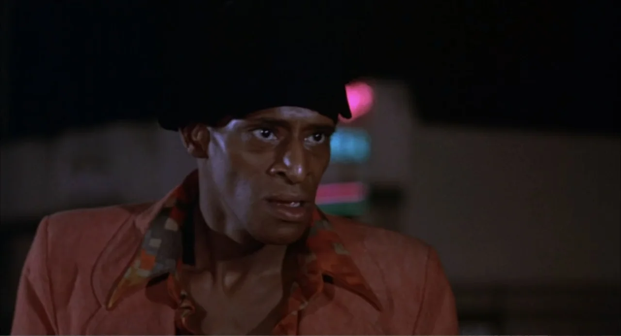 Foxy Brown (1974) movie still: medium — A dimly lit indoor space with soft ambient light hitting the subject. A man in a collared jacket looks…