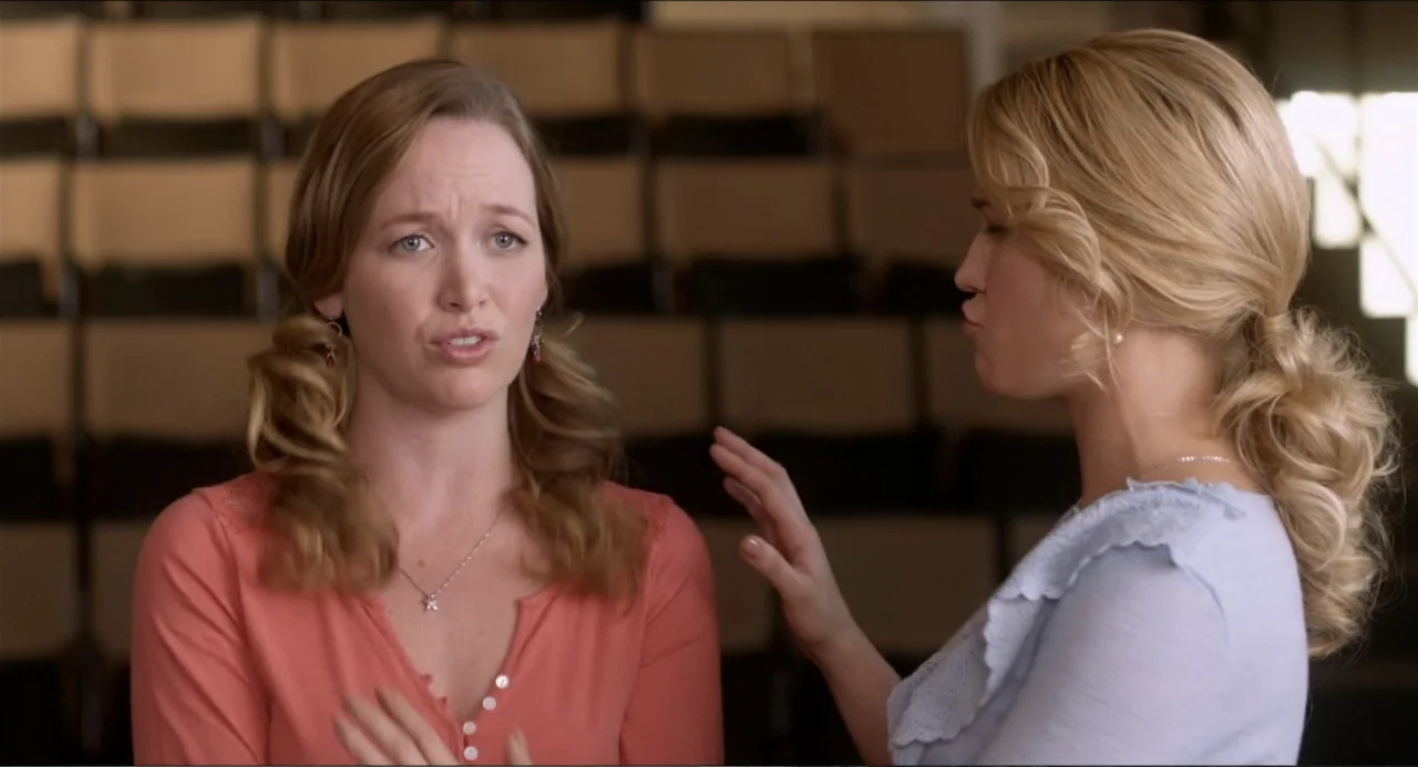 Pitch Perfect (2012) movie still: medium — An auditorium interior with rows of empty seats blurred in the background. Two young women stand in the…