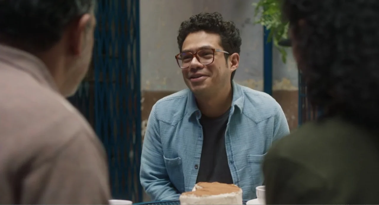 Casando a mi Ex (2023) movie still: medium — A cafe terrace features a metal table with a partially eaten dessert cake. Three people sit together in…