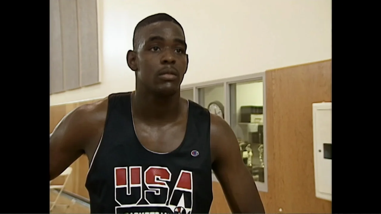 We Beat the Dream Team (2025) movie still: medium — An indoor basketball facility has tan walls and large windows looking into an office. A young…