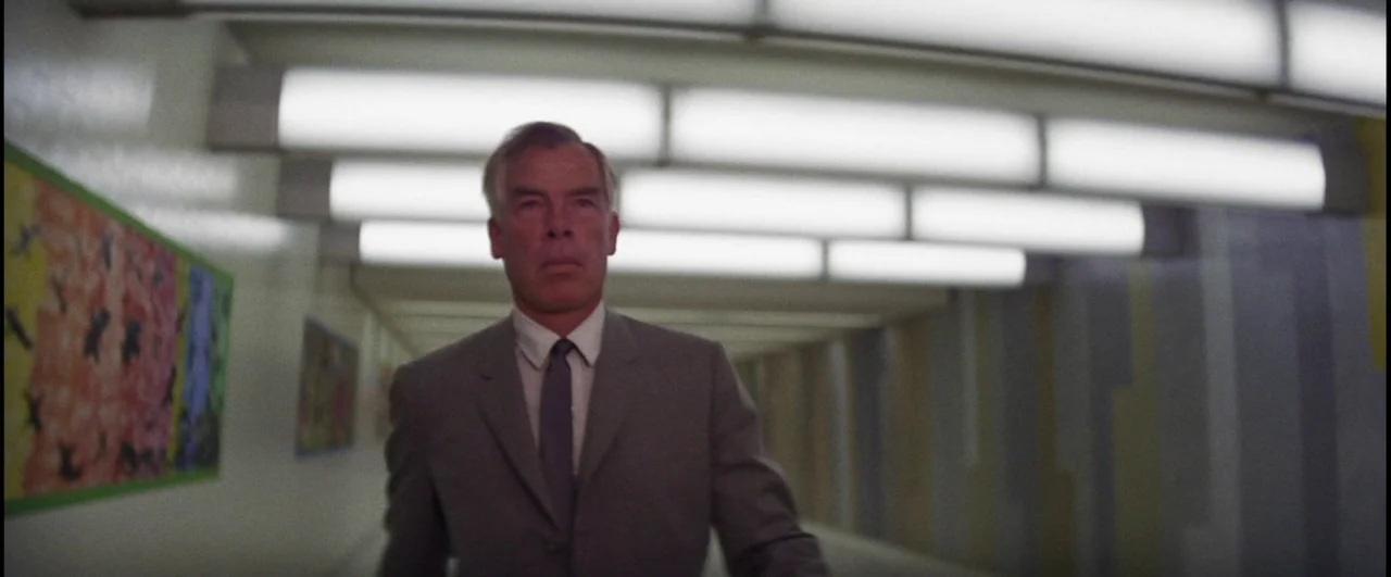 Point Blank (1967) movie still: medium — A long, white tiled pedestrian tunnel featuring modern art posters on the wall. A middle-aged man in a grey…