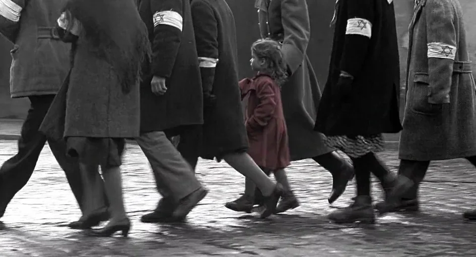 Schindler's List (1993) movie still: fullbody — A group of people walk together across a wet cobblestone street during the day. A small child…