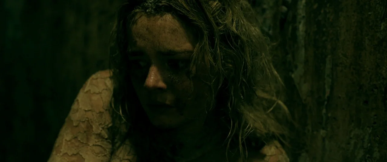 Ready or Not (2019) movie still: closeup — A dark, cramped stone tunnel with textured walls and faint grime. A young woman with disheveled hair and…