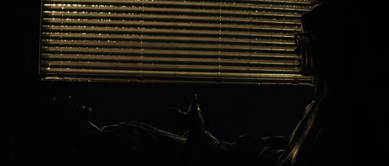 Suicide Squad (2016) movie still: medium — A dark room features a window covered by horizontal blinds at night. One person sits upright in profile…