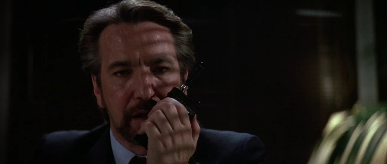 Die Hard (1988) movie still: closeup — A man speaks into a handheld radio within a dark, sparsely lit professional office. He faces slightly away…