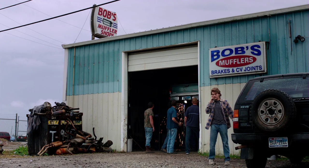 Killer Joe (2011) movie still: wide — A single-story metal building functions as a muffler shop under an overcast sky. Several men congregate…