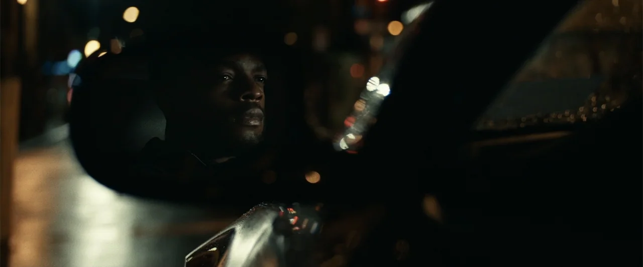 21 Bridges (2019) movie still: closeup — A man's face is reflected in the side mirror of a dark car at night. The man sits in the driver seat…