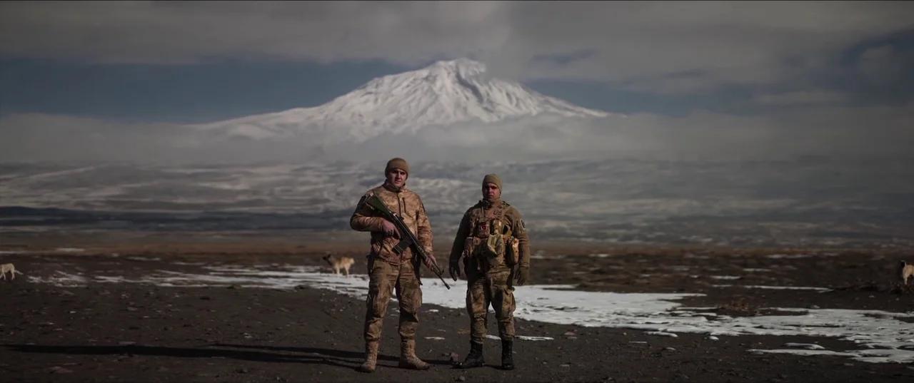 About Dry Grasses (2023) movie still: fullbody — Two soldiers in camouflage stand on a vast high-altitude plateau with patches of snow. A massive…