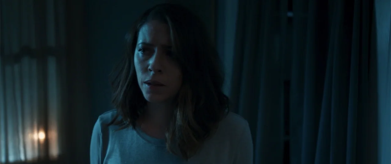 Phobias (2021) movie still: medium — A dimly lit room at night features heavy curtains covering a window. A woman stands in the center of the frame…