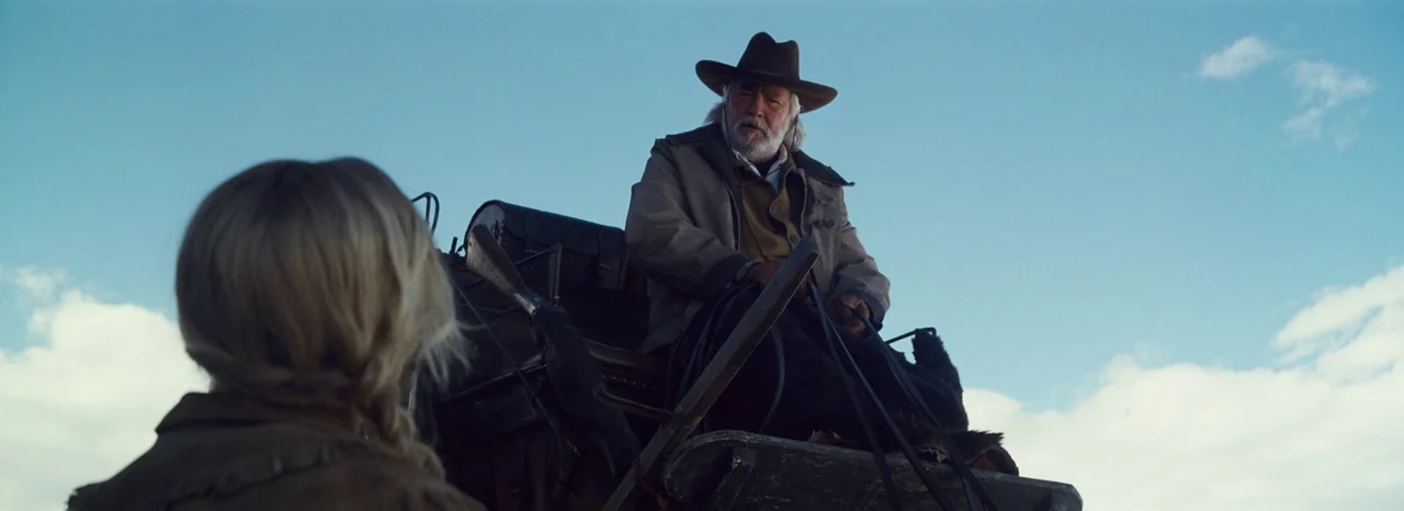 The Hateful Eight (2015) movie still: medium — An open landscape with a clear blue sky serves as the backdrop. A bearded older man in a brown hat…