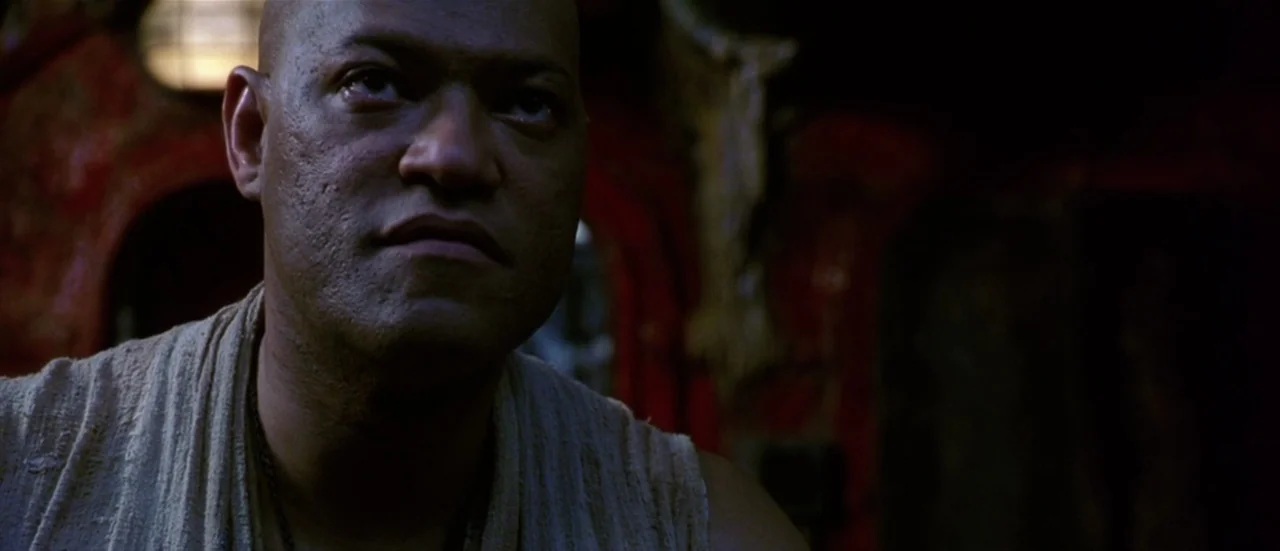 The Matrix Reloaded (2003) movie still: closeup — A dimly lit interior space with textured red metal walls and mechanical details. A bald man stares…