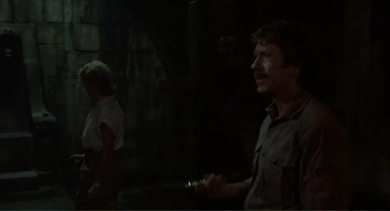 Firewalker (1986) movie still: medium — A dark, ancient stone chamber features heavy masonry walls and deep shadows. A man holding an object stands…
