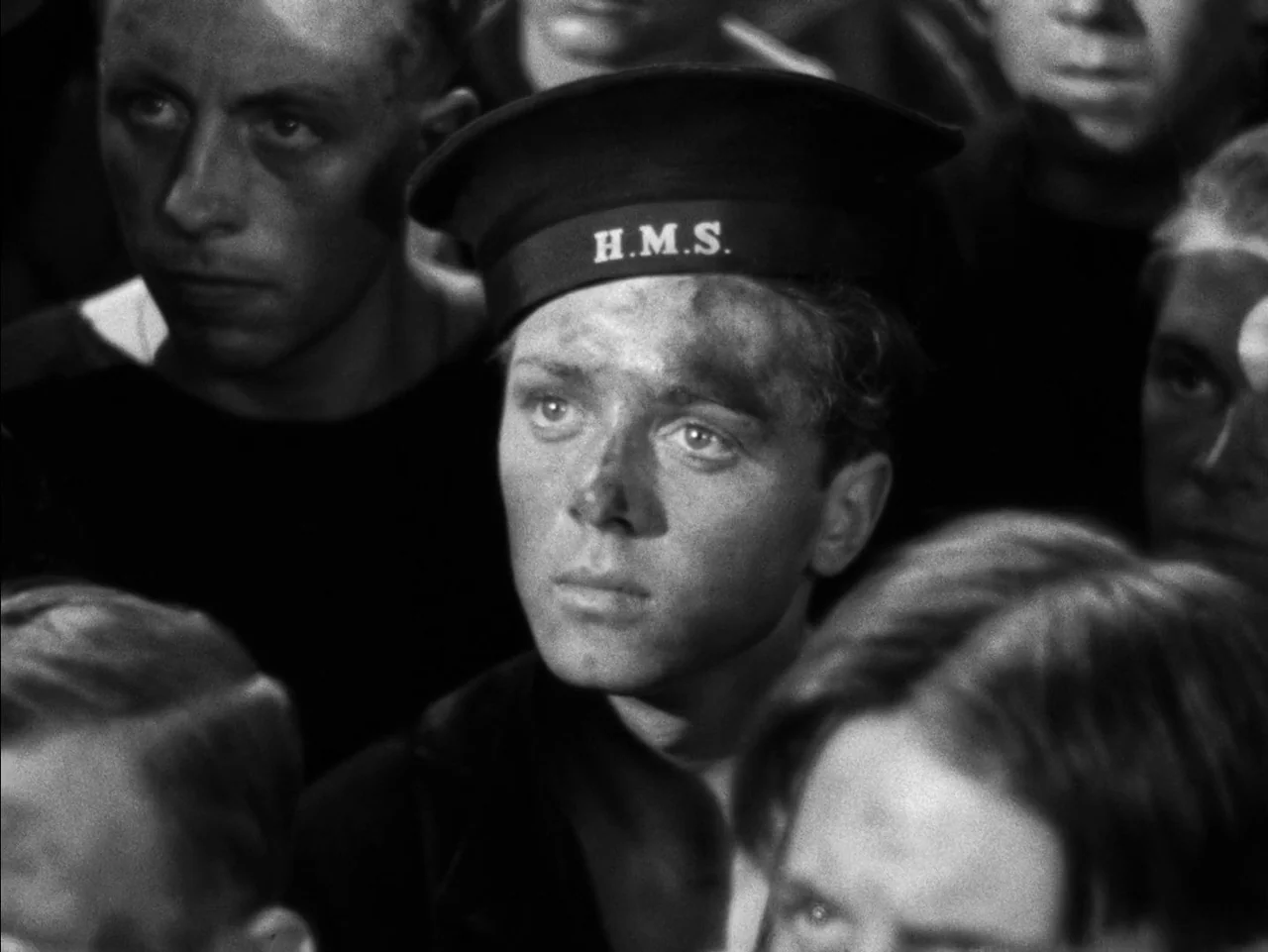In Which We Serve (1942) movie still: closeup — A close-up of a young sailor on a ship with others in the background. The sailor has soot on his…