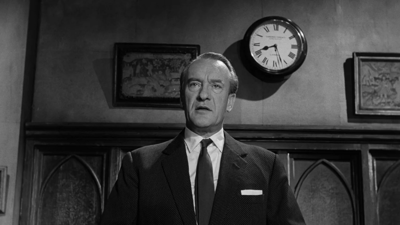 Village of the Damned (1960) movie still: medium — A man stands in front of a wood-paneled wall with framed pictures and a clock. The man looks…