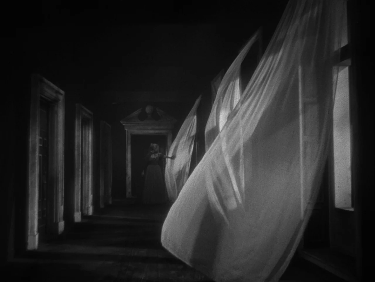 La Belle et la Bête (1946) movie still: wide — A dark, empty corridor features multiple doorways and tall windows along the side wall. Large white…
