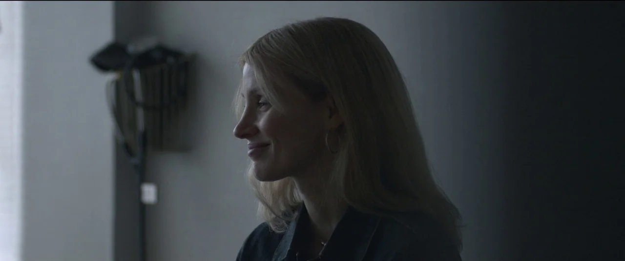 The Good Nurse (2022) movie still: medium — A dimly lit room with plain walls features a standing floor lamp. A blonde woman in a denim jacket is…