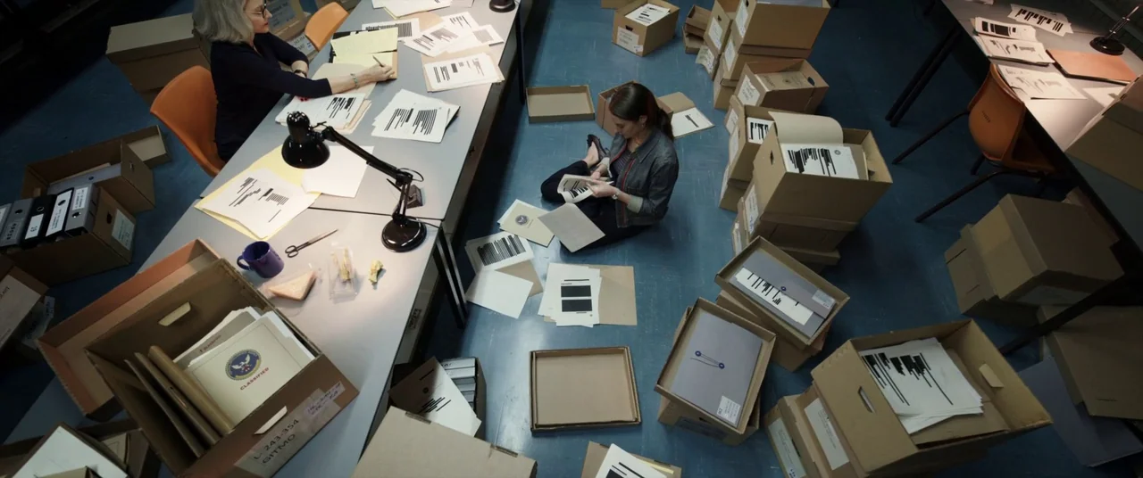 The Mauritanian (2021) movie still: wide — A cluttered archive room contains desks and dozens of cardboard boxes filled with papers. A younger woman…