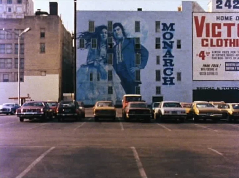 Mur Murs (1981) movie still: wide — A paved parking lot sits before a multi-story building with a large mural painted. The mural depicts a man and…