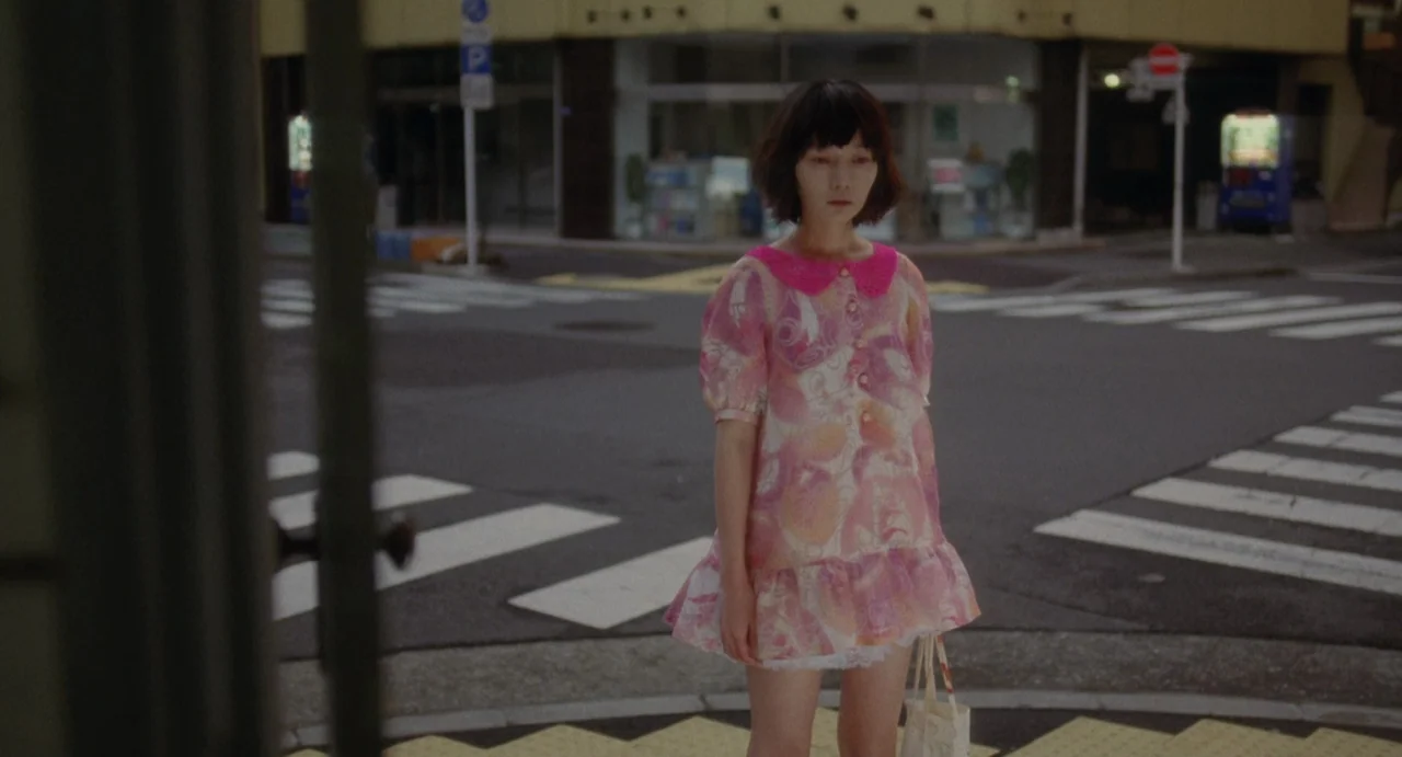 Air Doll (2009) movie still: fullbody — An urban intersection with painted crosswalks and a nearby vending machine. A young woman stands alone on…