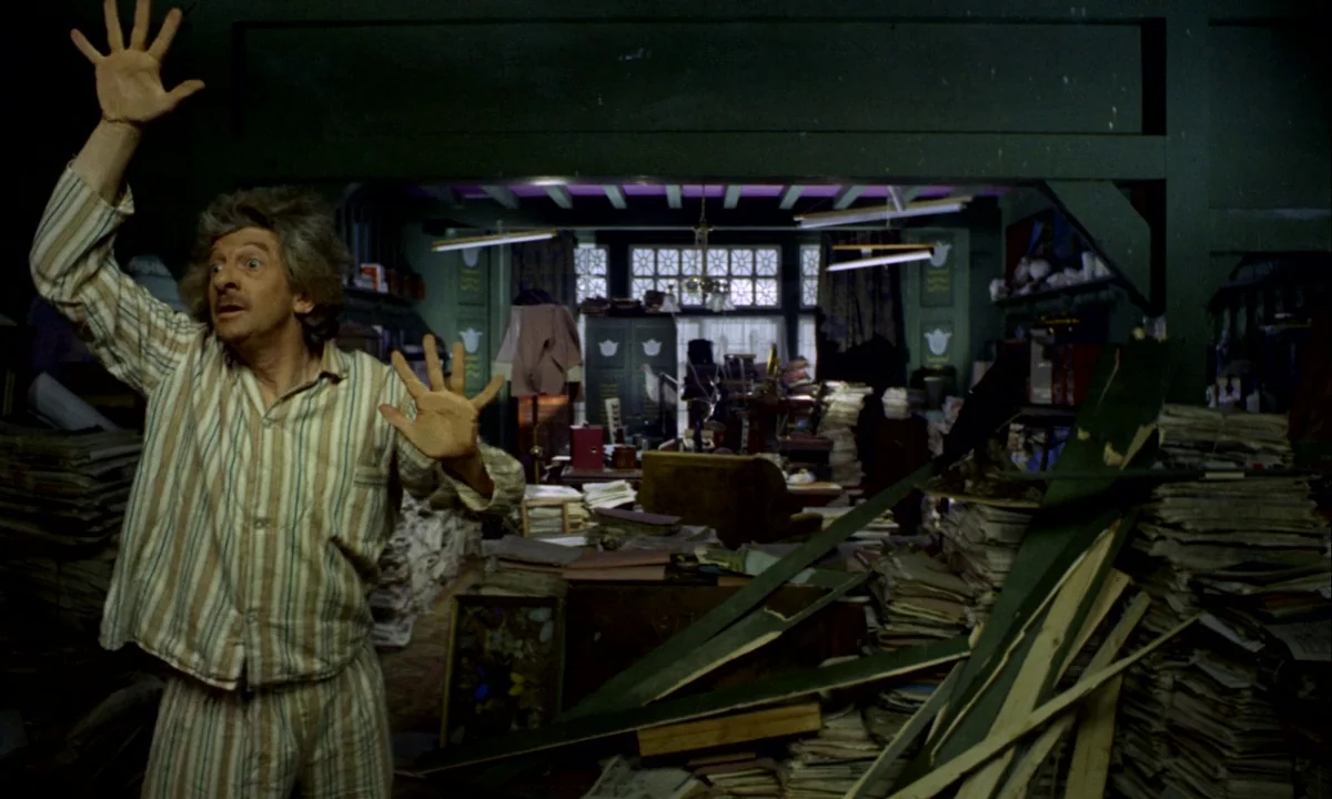 Wonderwall (1968) movie still: medium — A dark cluttered room is filled with stacks of paper and broken wooden debris. A man in pajamas stands with…