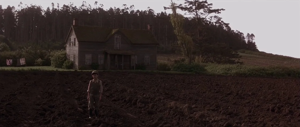 Snow Falling on Cedars (1999) movie still: wide — A weathered wooden farmhouse sits at the edge of a vast, dark plowed field. A young boy stands…