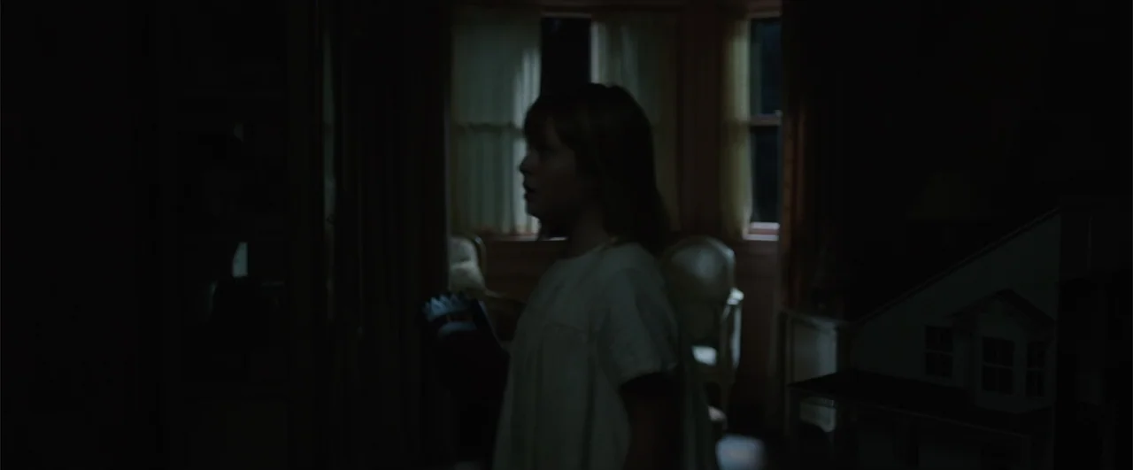 Annabelle: Creation (2017) movie still: medium — A dimly lit room contains furniture and a toy house in the shadows. A child with shoulder-length…