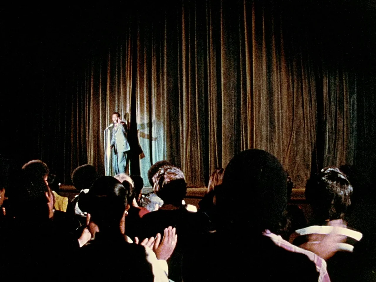 Space is the Place (1974) movie still: wide — A darkened auditorium with a silhouetted audience facing a lit stage with curtains. A lone man in a…