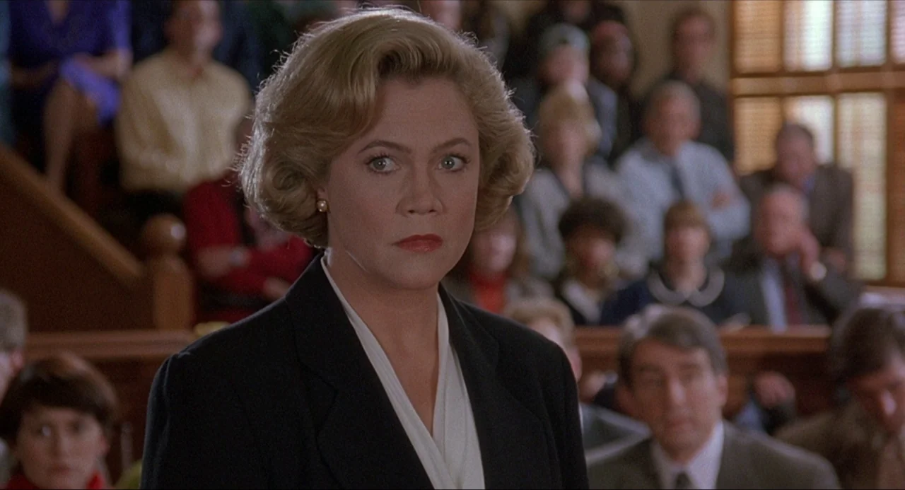 Serial Mom (1994) movie still: medium — A courtroom filled with blurred observers sitting in the background benches. A woman in a dark blazer stands…