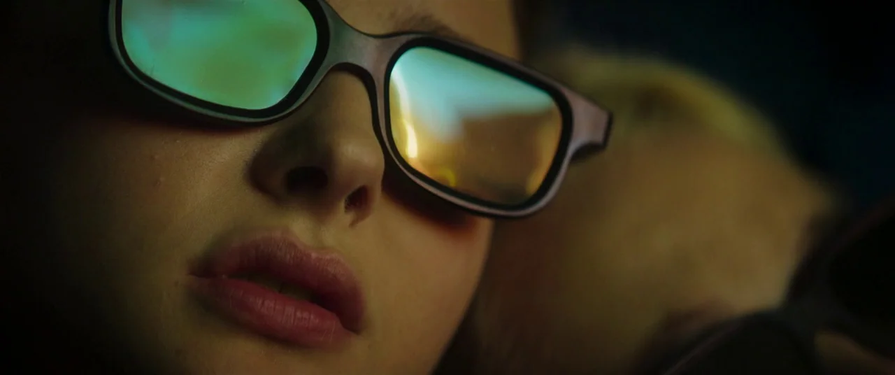 Greta movie still: closeup — A close view shows the lower face of a person wearing plastic 3D glasses. Light from an unseen movie screen reflects on…