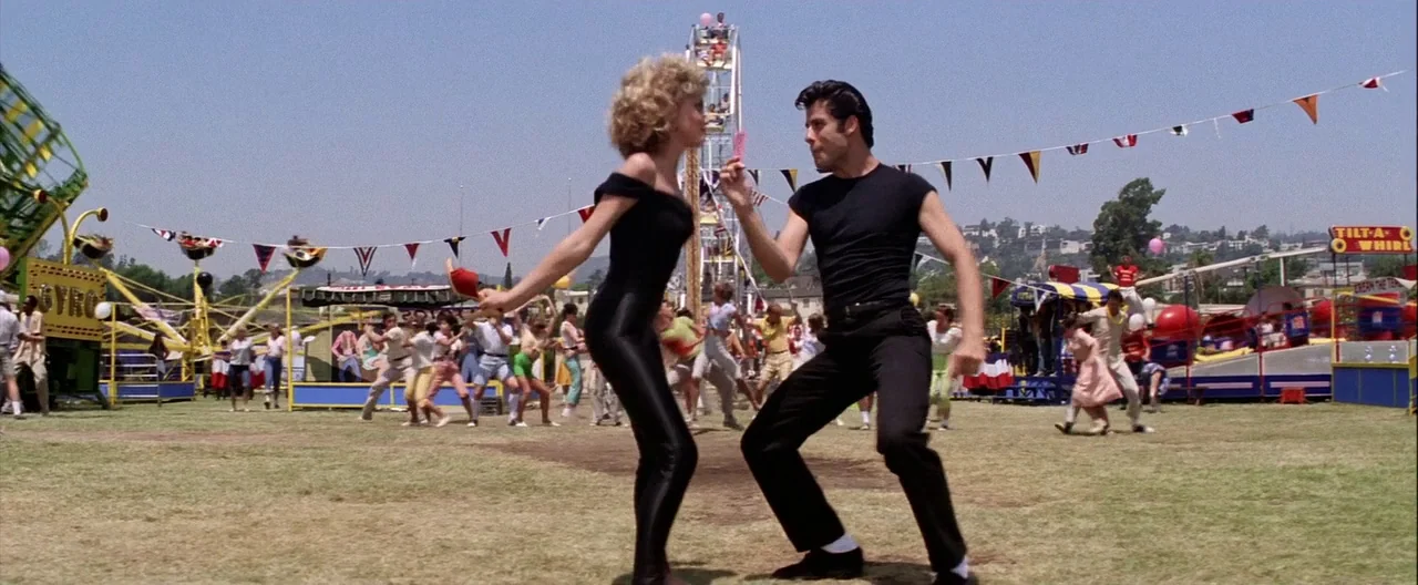 Grease (1978) movie still: fullbody — A carnival fairground features various rides and colorful bunting under a clear sky. A man and woman in black…