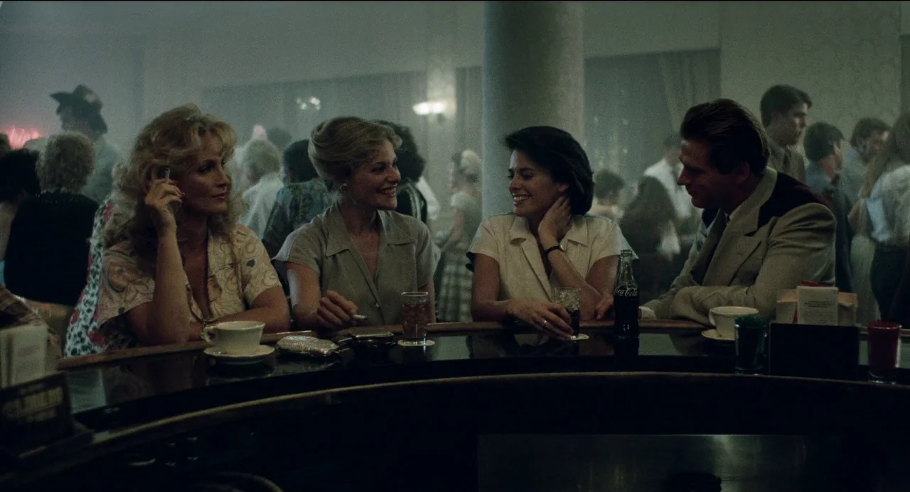 Desert Hearts (1985) movie still: medium — A crowded bar interior with low light and hazy atmosphere featuring a curved counter. Four people sitting…
