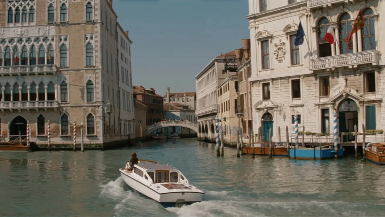 The Tourist (2010) movie still: wide — A Venetian canal is flanked by tall ornate historic stone buildings with arched windows. A white motorboat…