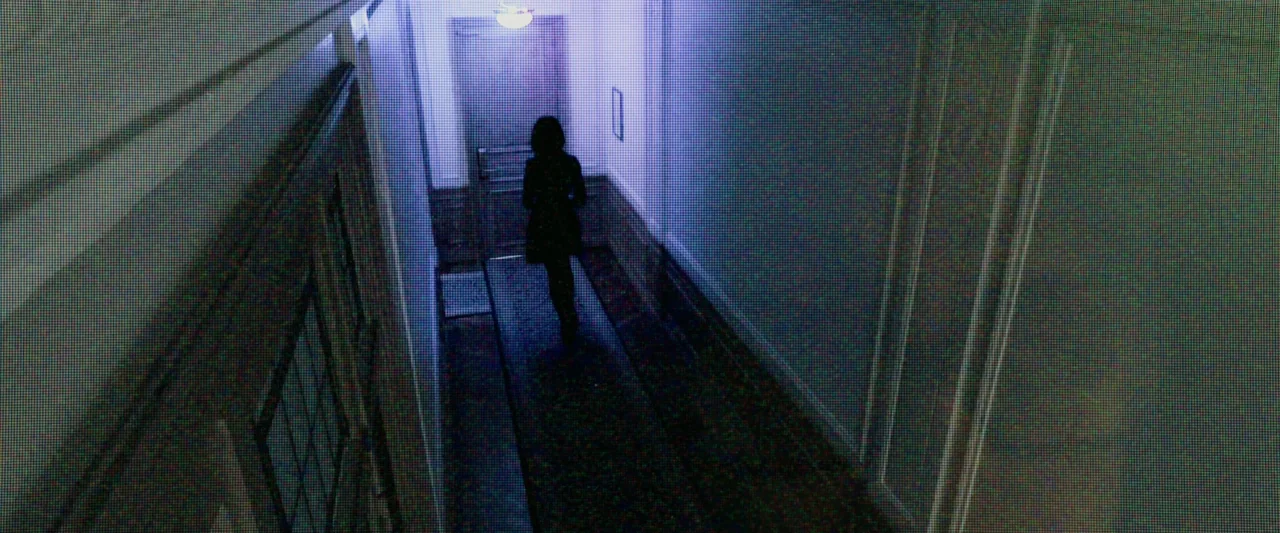 Anna (2019) movie still: wide — A long, narrow hallway features high walls and a single door at the end. A lone individual walks away from the…